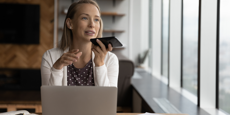 Female read person with long blond hair and white cardigan talking into a phone while using a laptop